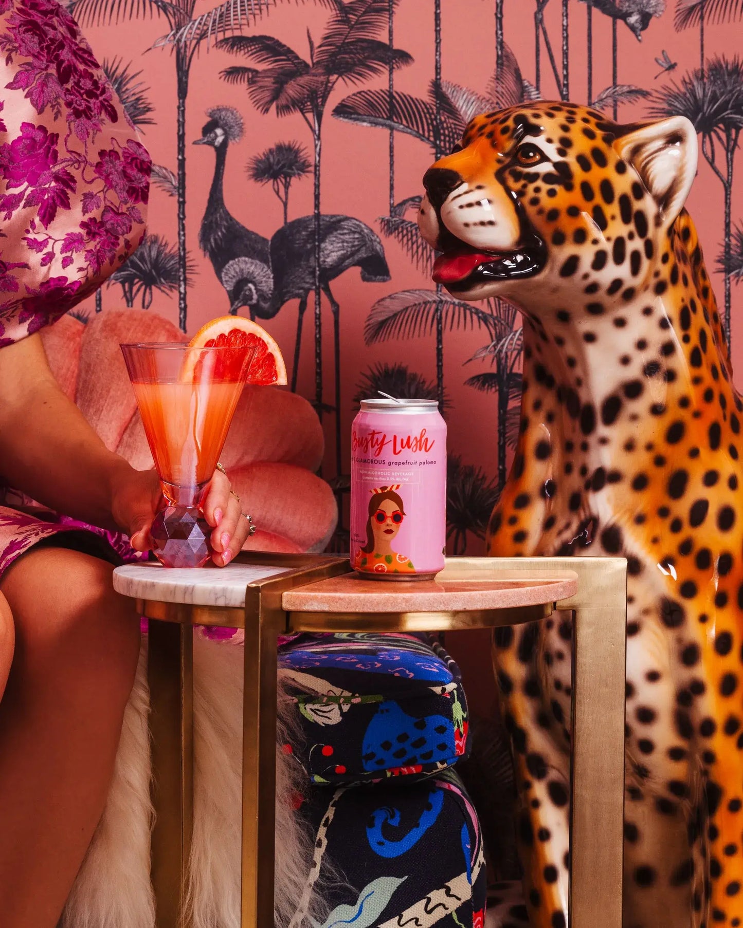 Person sitting with a Busty Lush Grapefruit Paloma in a Glass and can, leopard statue, and colorful background.